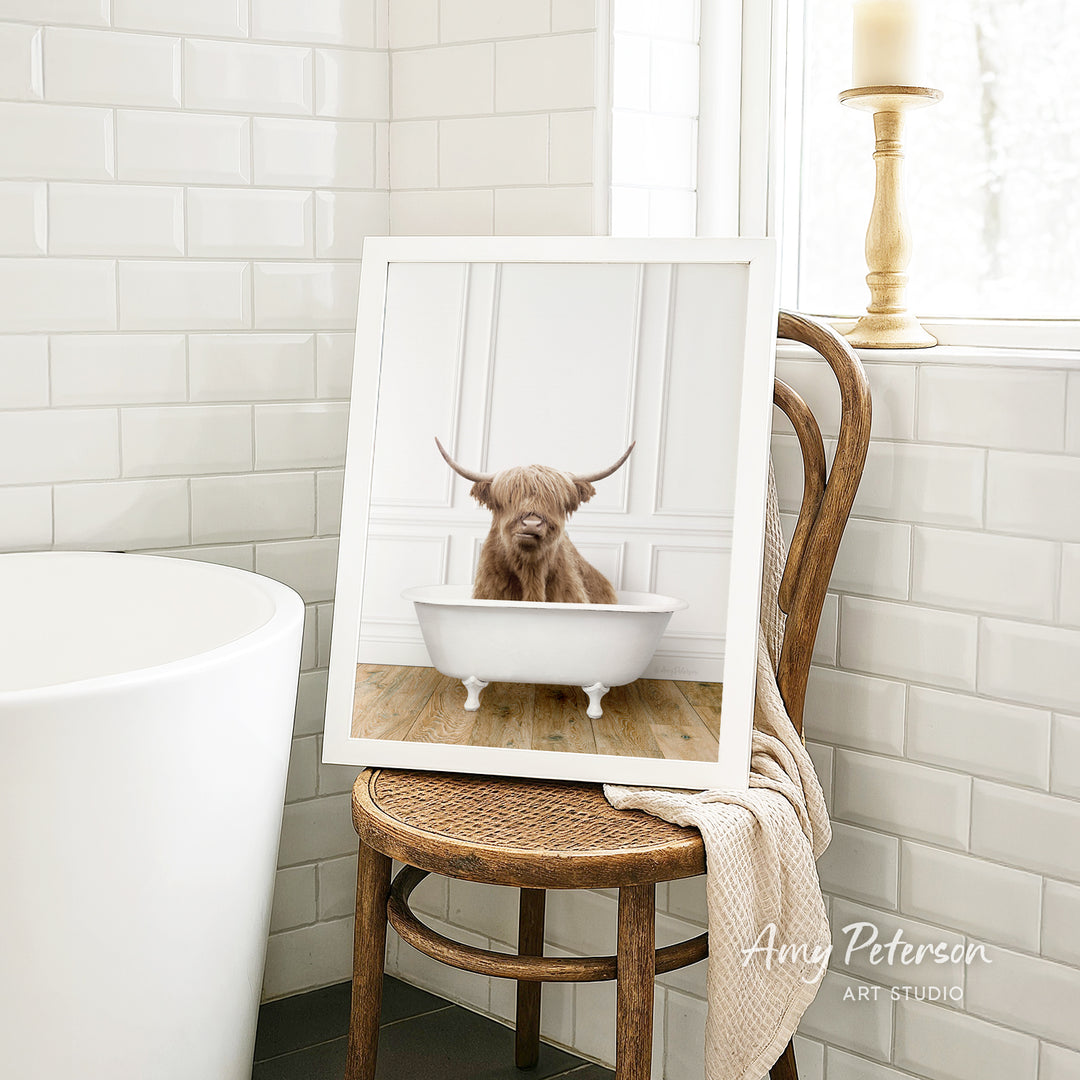 A framed image of a Highland cow sitting in a white bathtub, with a wooden chair and a towel in the background.