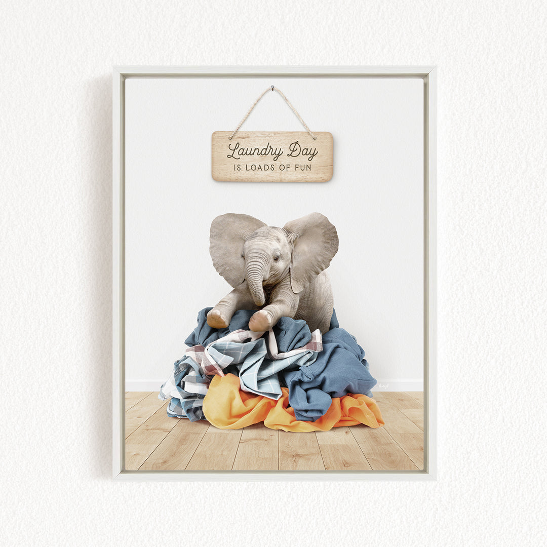 A framed image of a stuffed elephant sitting atop a pile of laundry, with a wooden sign above that reads "Laundry Day is loads of fun".
