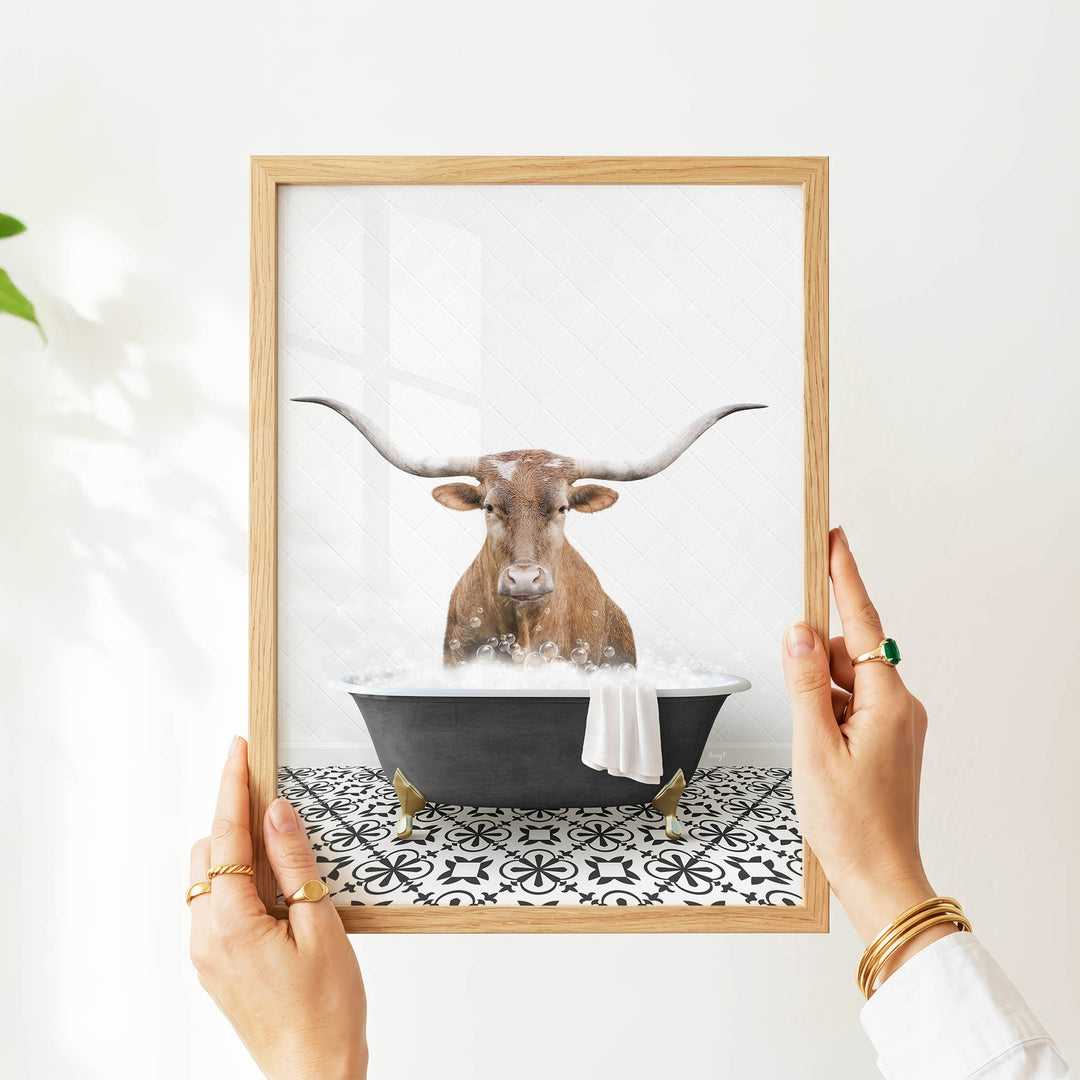 A person's hands are holding a framed picture of a bull with long horns, which is sitting in a bathtub filled with water and bubbles.