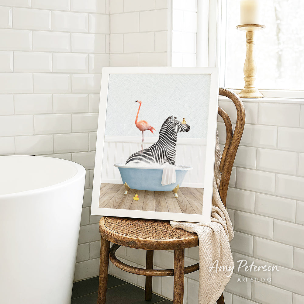 A framed painting of a zebra sitting in a bathtub, with a pink flamingo standing on its back, is displayed on a wooden chair in a bathroom setting.