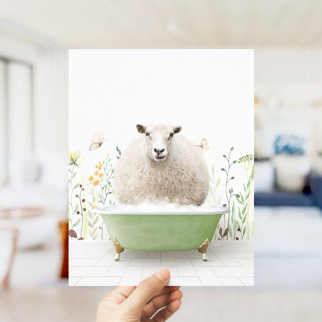 A hand holds up a framed image depicting a fluffy white sheep sitting in a green bathtub filled with bubbles, surrounded by a floral pattern.
