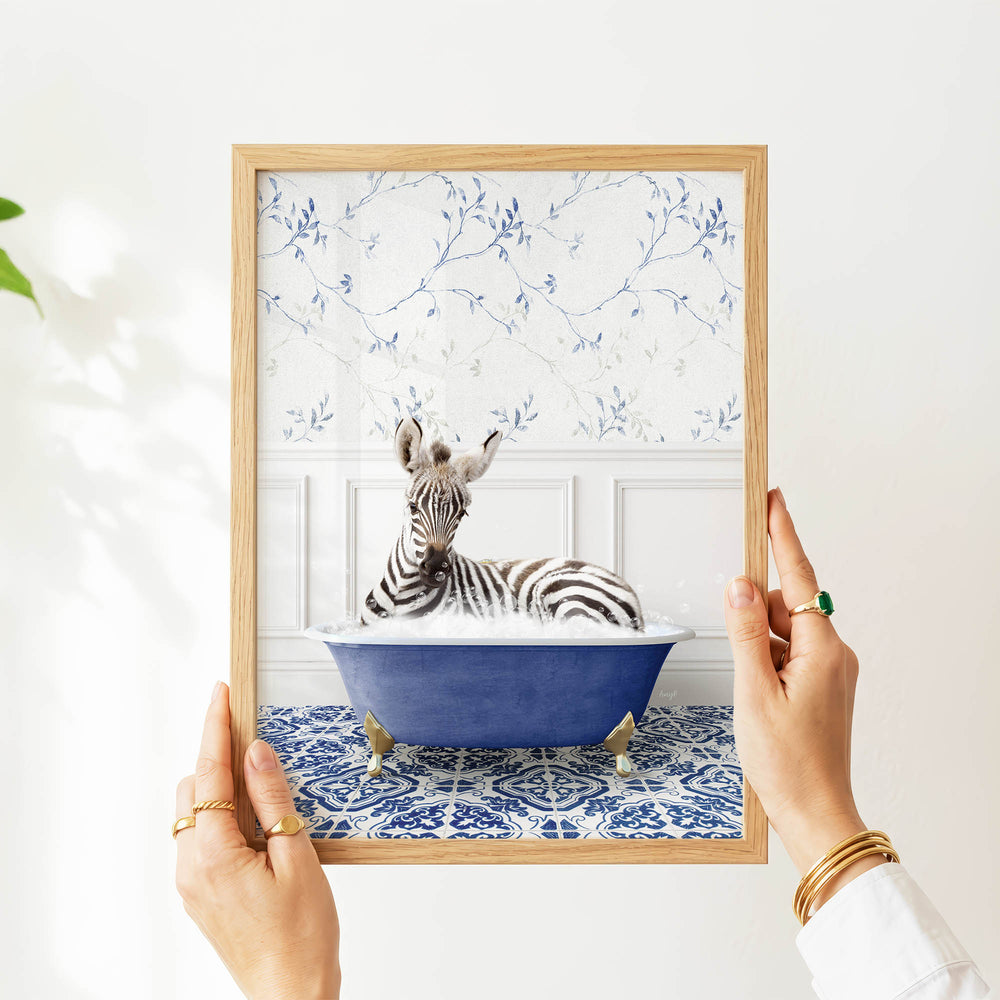 A person's hands are holding a framed picture of a zebra sitting in a blue bathtub, with a patterned wall behind it.