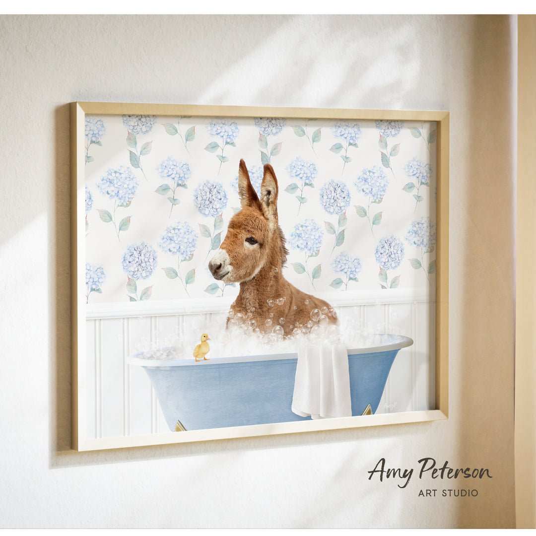 A donkey is sitting in a blue bathtub filled with bubbles, with a yellow duck nearby, against a backdrop of blue hydrangea flowers.