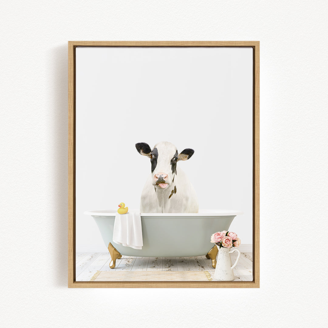 A black and white cow is sitting in a bathtub filled with water, surrounded by a yellow rubber duck, a white towel, and a vase of pink flowers.