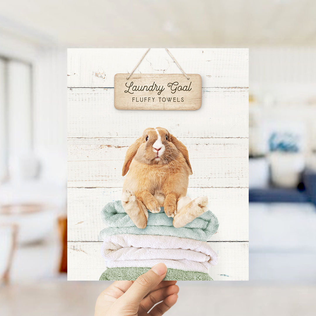 A hand holding a card with a picture of a rabbit sitting on a stack of fluffy towels, with a wooden sign above it that says "Laundry Goal Fluffy Towels".