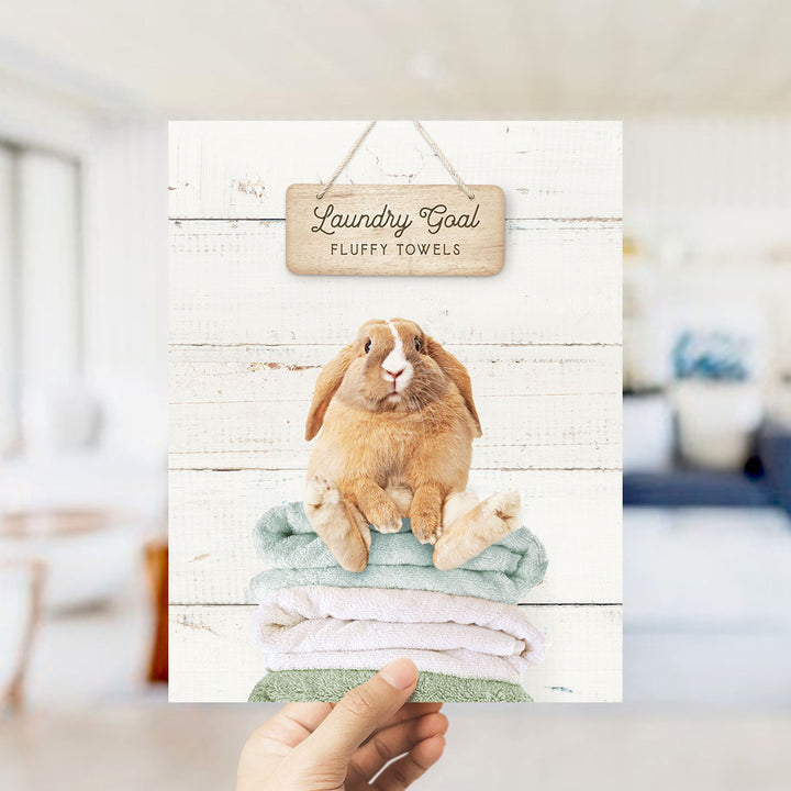 A hand holding a card with a picture of a rabbit sitting on a stack of fluffy towels, with a wooden sign above it that says "Laundry Goal Fluffy Towels".