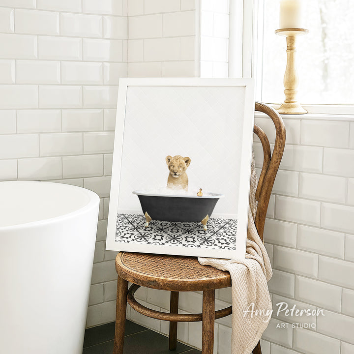 A framed image of a lion sitting in a bathtub, surrounded by a patterned tile floor, is displayed on a wooden chair in a bathroom setting.