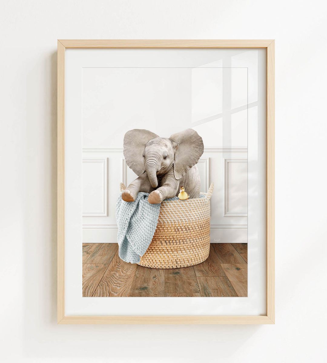 A framed photograph of a baby elephant sitting in a woven basket, with a blue blanket and a yellow toy nearby.