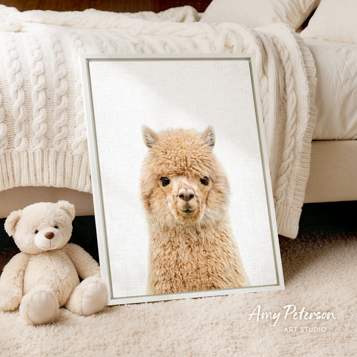 A framed image of a fluffy alpaca is displayed on a bed, accompanied by a teddy bear.