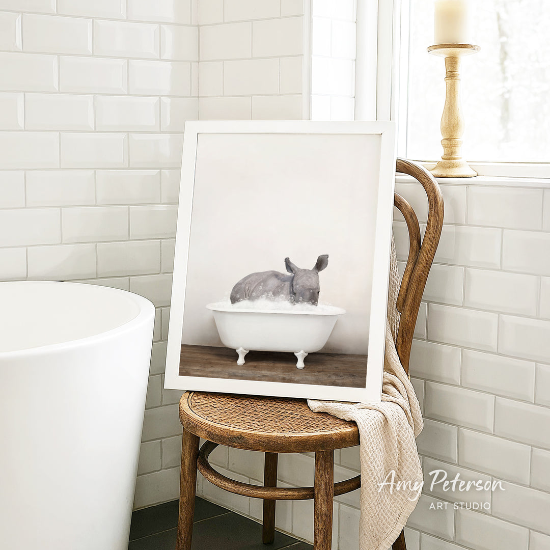 A framed photograph of a rhinoceros sitting in a bathtub is displayed on a wooden chair in a bathroom setting.