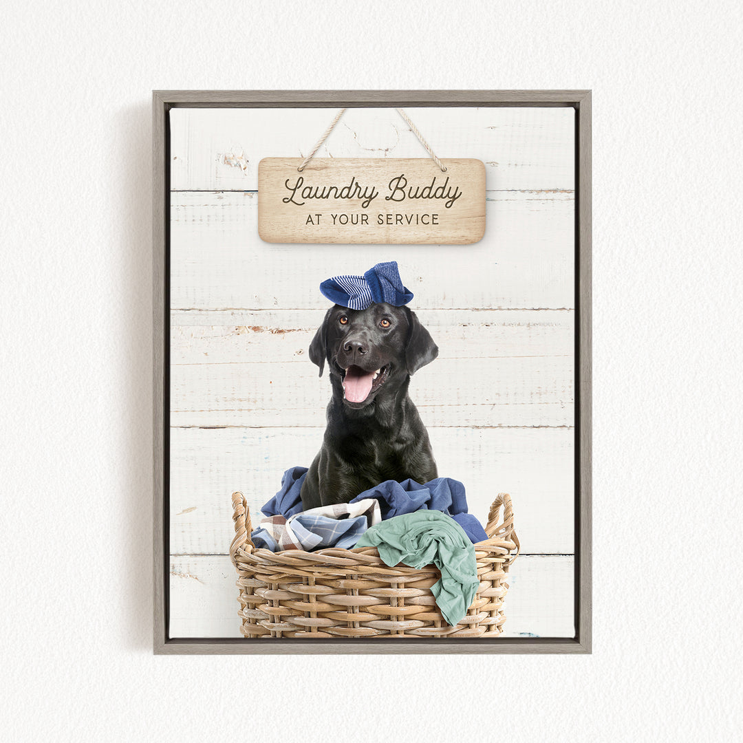 A black dog wearing a blue hat sits in a wicker basket filled with clothes, with a wooden sign above that reads "Laundry Buddy at your service".