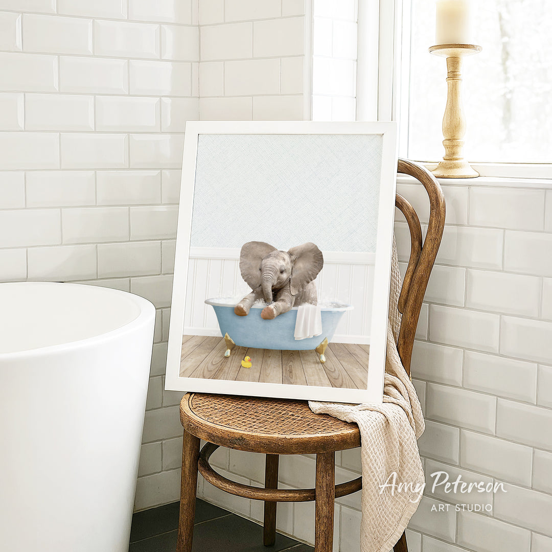 A framed image of a baby elephant sitting in a bathtub, with a towel draped over the side of the tub.