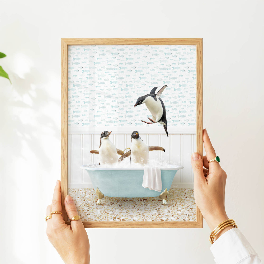 A person's hands are holding a framed picture that depicts two penguins sitting in a bathtub filled with bubbles, with one penguin jumping out of the tub.