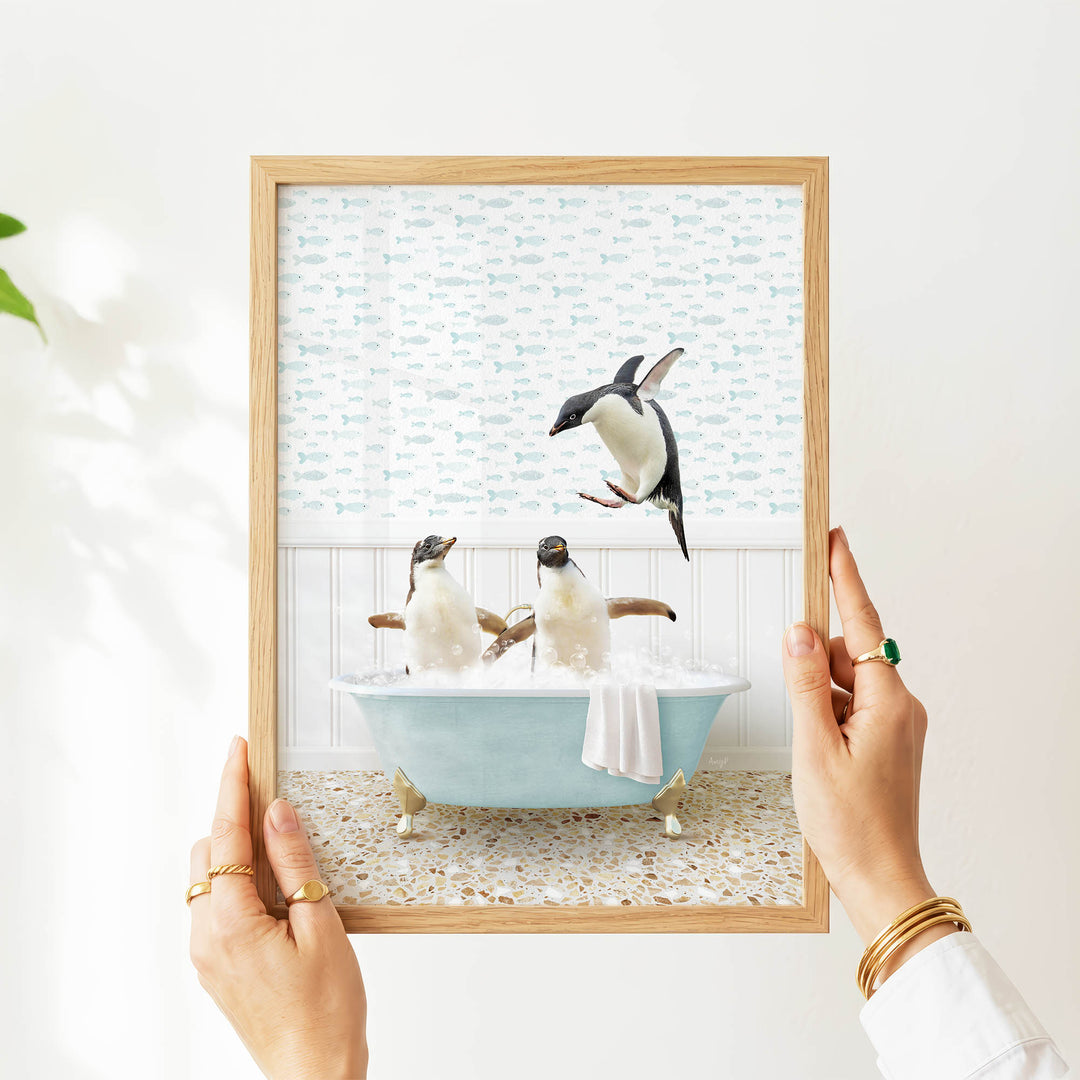 A person's hands are holding a framed picture that depicts two penguins sitting in a bathtub filled with bubbles, with one penguin jumping out of the tub.