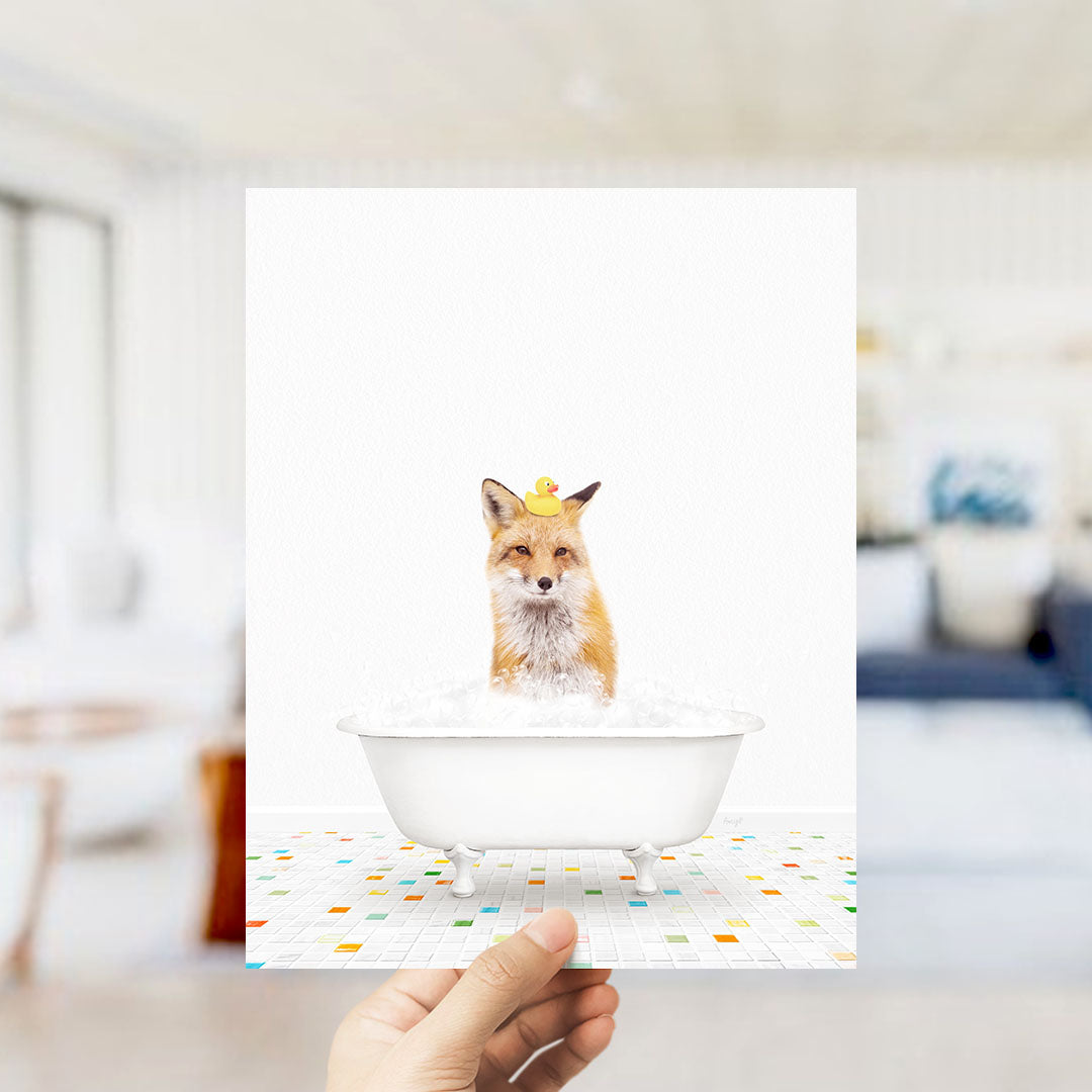 A hand holds up a framed image of a fox sitting in a bathtub filled with bubbles, with a yellow rubber duck perched on its head.