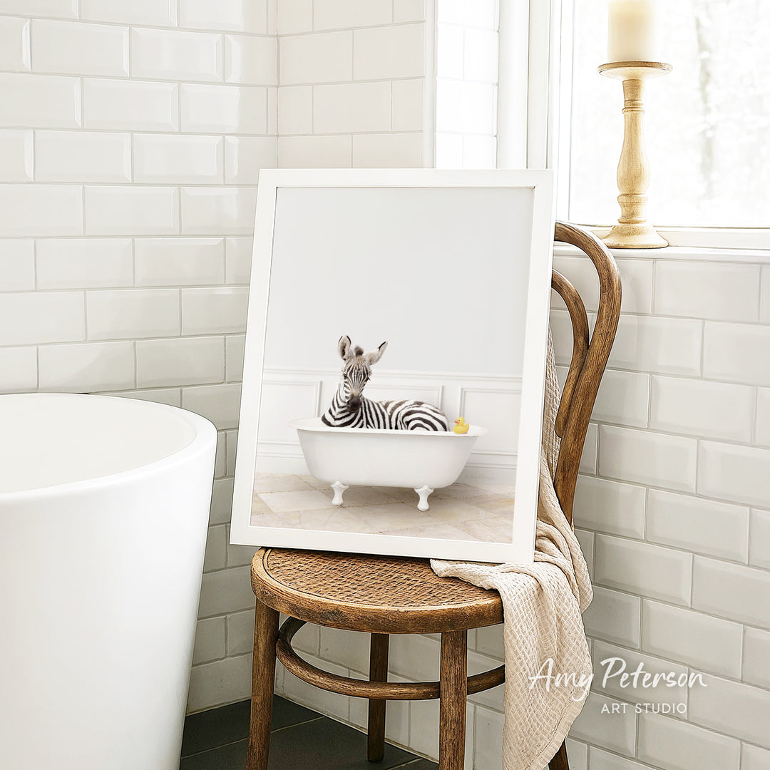 A framed image of a zebra sitting in a bathtub, with a towel draped over the back of a wooden chair in a bathroom setting.