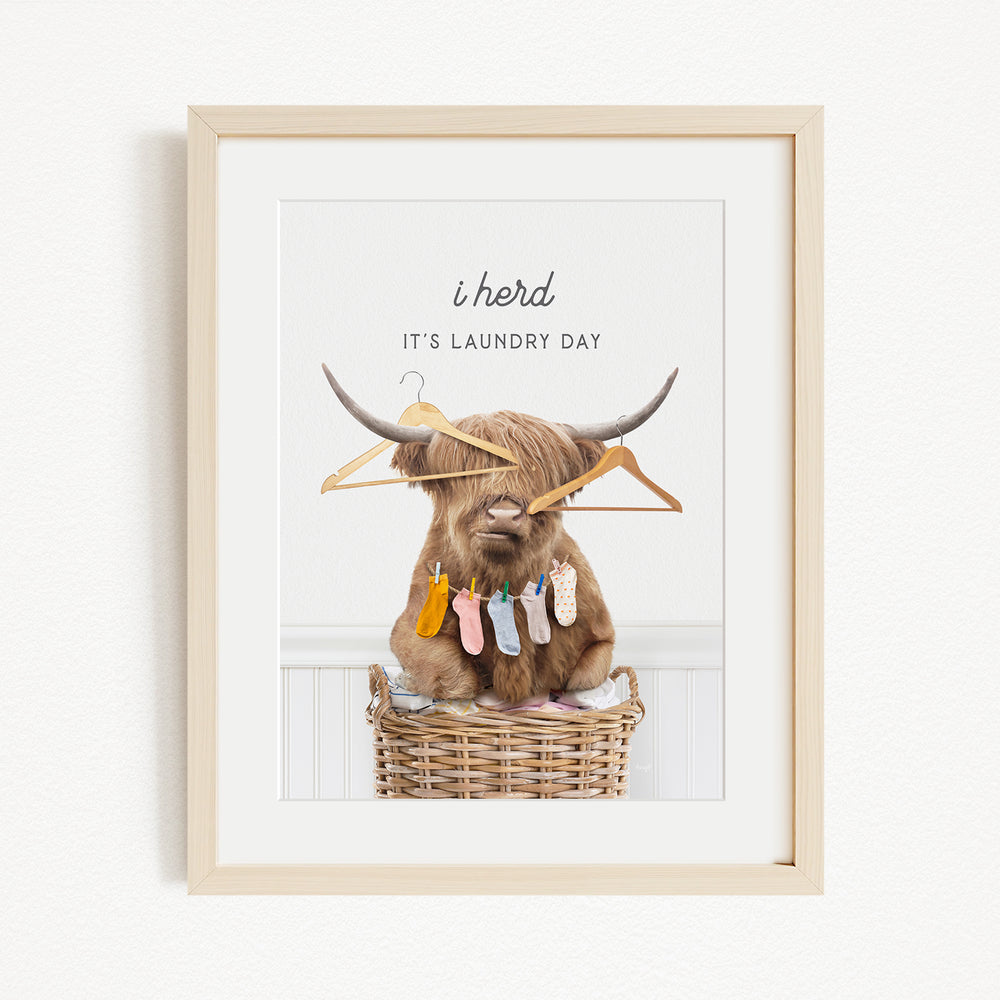 A framed image depicting a brown cow with large horns sitting in a wicker basket, surrounded by clothes and hangers, with the text "It's laundry day" above the cow.