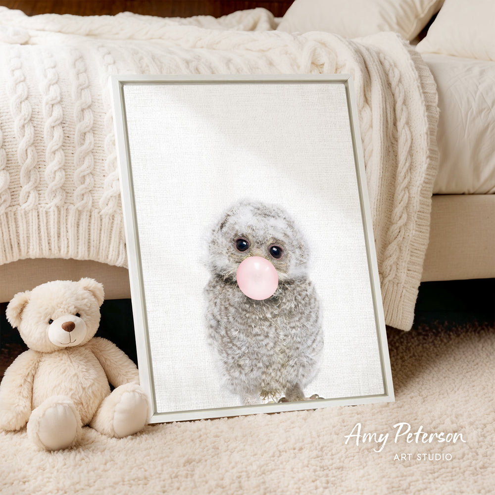 A framed print of a gray owl blowing a bubble gum bubble is displayed on a beige blanket, with a teddy bear sitting next to it on a beige carpet.