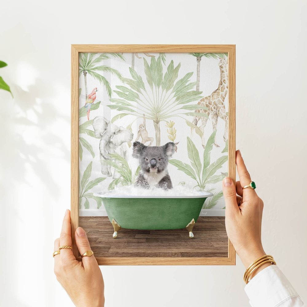 A person's hands are holding a framed picture of a koala sitting in a green bathtub, surrounded by tropical foliage.