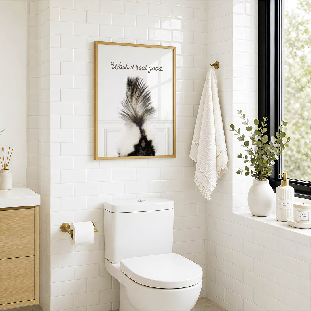 A modern bathroom with a white toilet, a framed poster of a skunk, and a window with greenery.