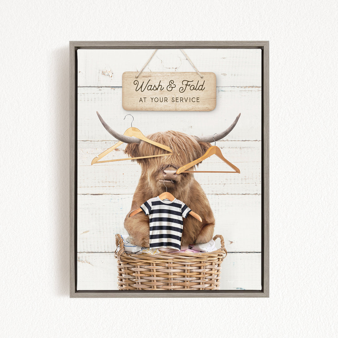 A framed image depicting a large, furry animal, possibly a yak or a yak dog, sitting in a wicker basket and holding a striped shirt. The animal is surrounded by clothes hangers, and there is a sign above the basket that reads "Wash & Fold at your Service".