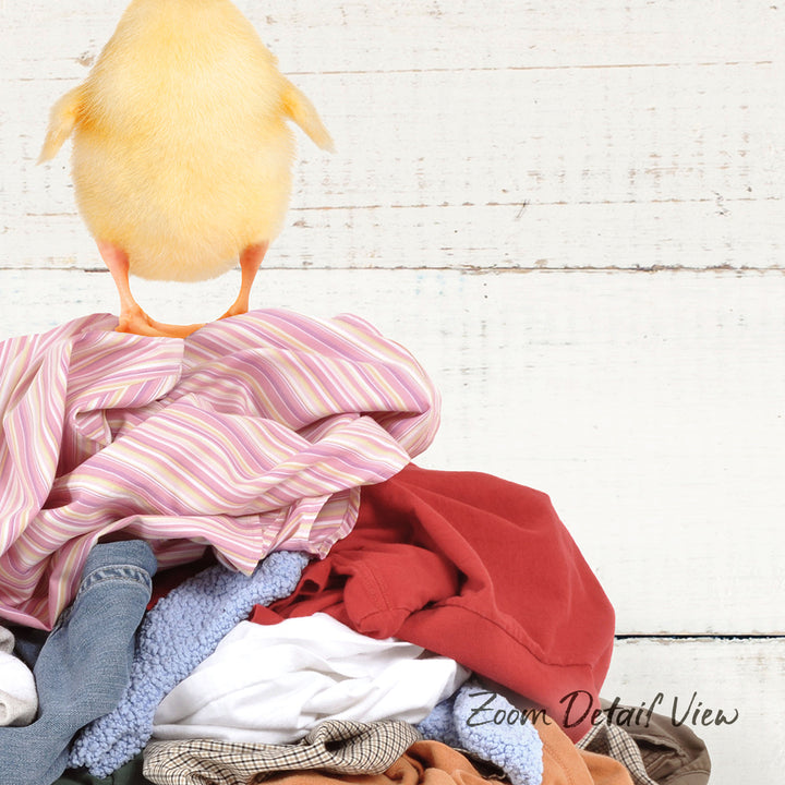 A yellow chick stands atop a pile of clothes on a white wooden surface.