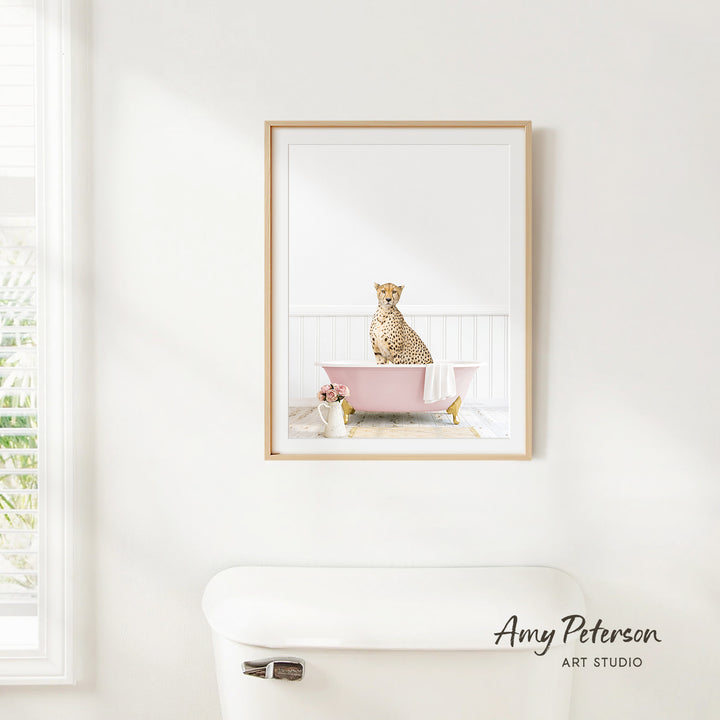 A framed image of a cheetah sitting in a pink bathtub is hanging on a white wall in a bathroom.