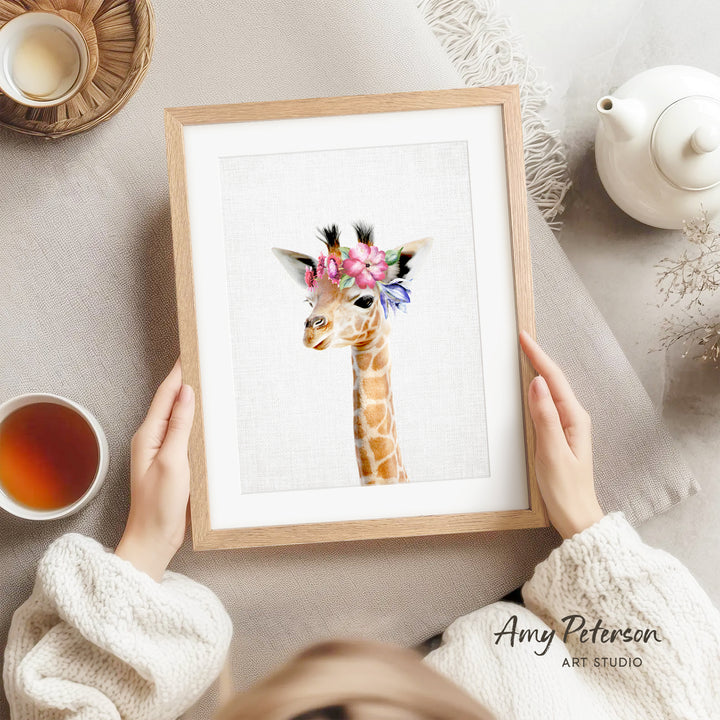 A person is holding a framed print of a giraffe with a floral headpiece, surrounded by various objects such as a teapot, a cup, and a bowl.