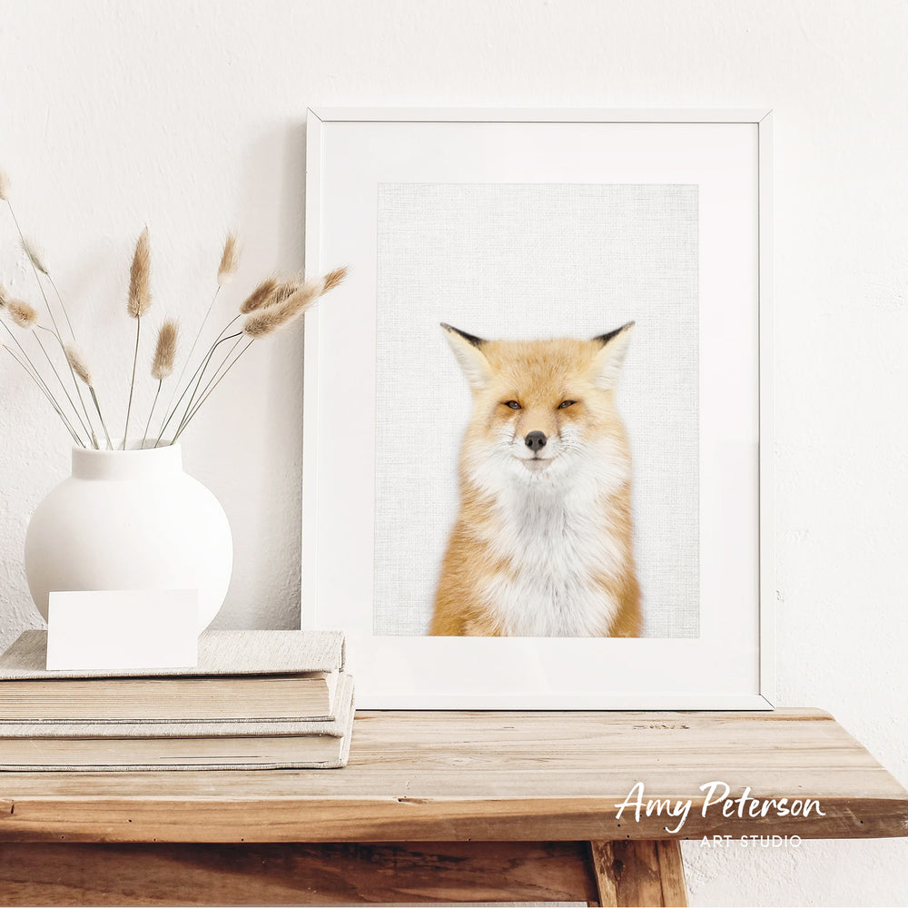 a framed picture of a fox with a white background, placed on a wooden shelf next to a white vase with dried flowers.