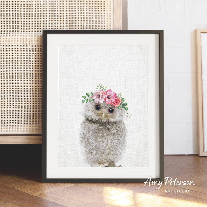 A framed print of a cute owl wearing a flower crown is displayed on a wooden floor.