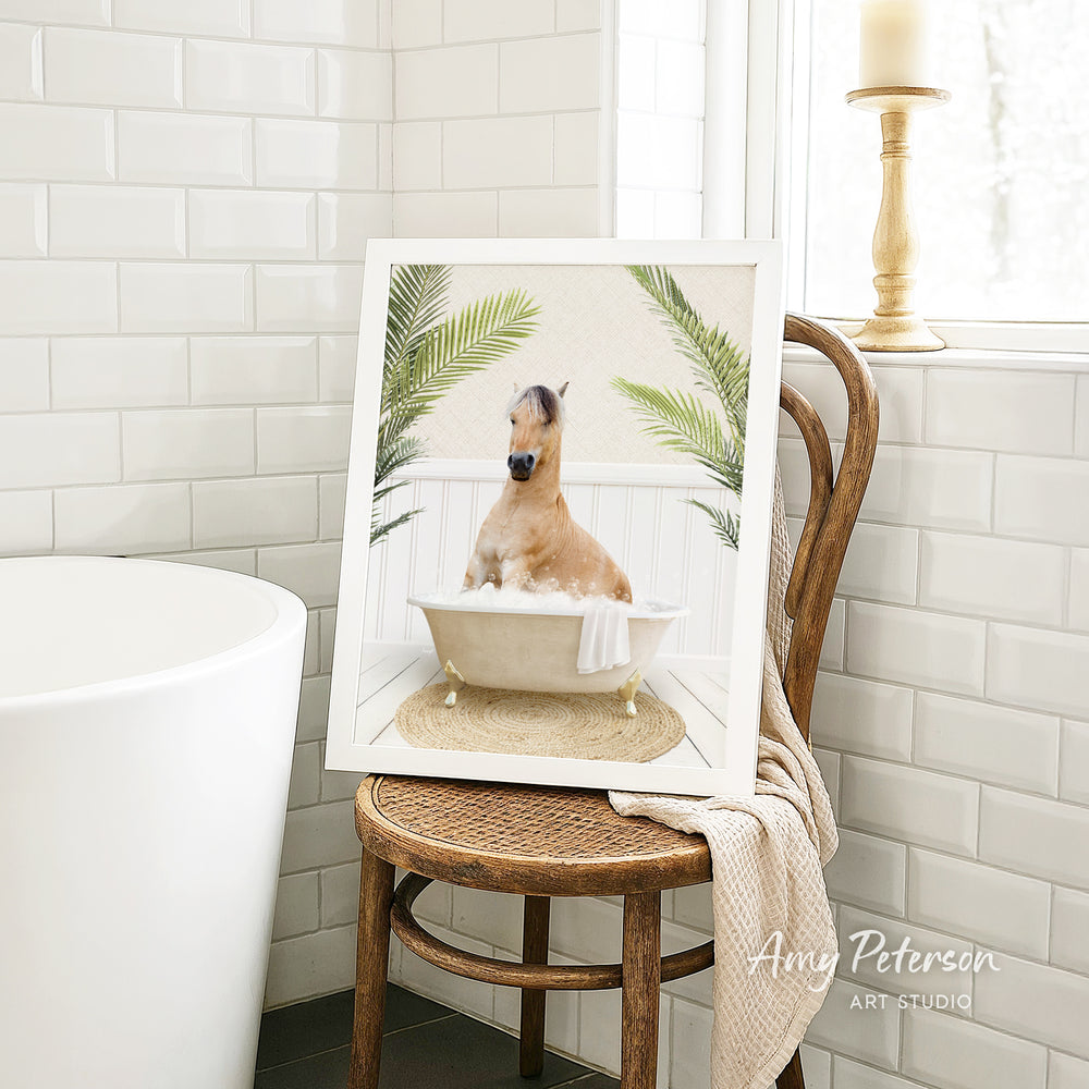 A framed image of a horse sitting in a bathtub, surrounded by palm leaves, is displayed on a wooden chair in a bathroom setting.