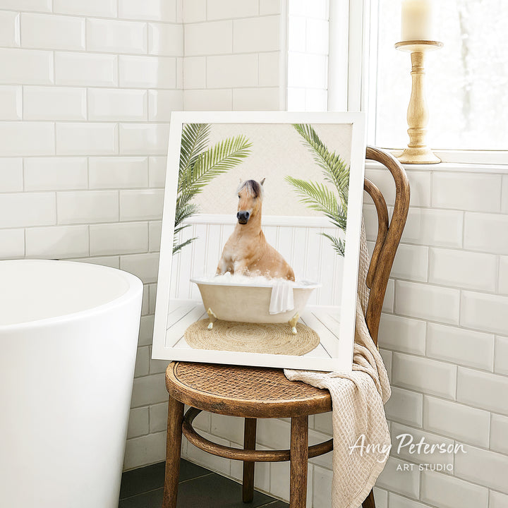 A framed image of a horse sitting in a bathtub, surrounded by palm leaves, is displayed on a wooden chair in a bathroom setting.