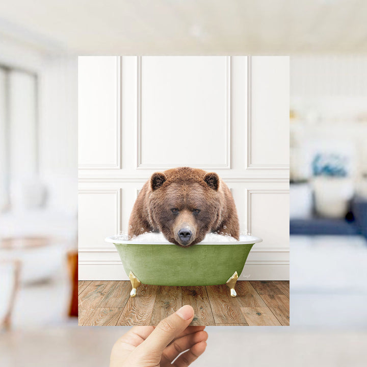 A hand holds up a photo of a brown bear sitting in a green bathtub, with a blurred background suggesting a bathroom setting.
