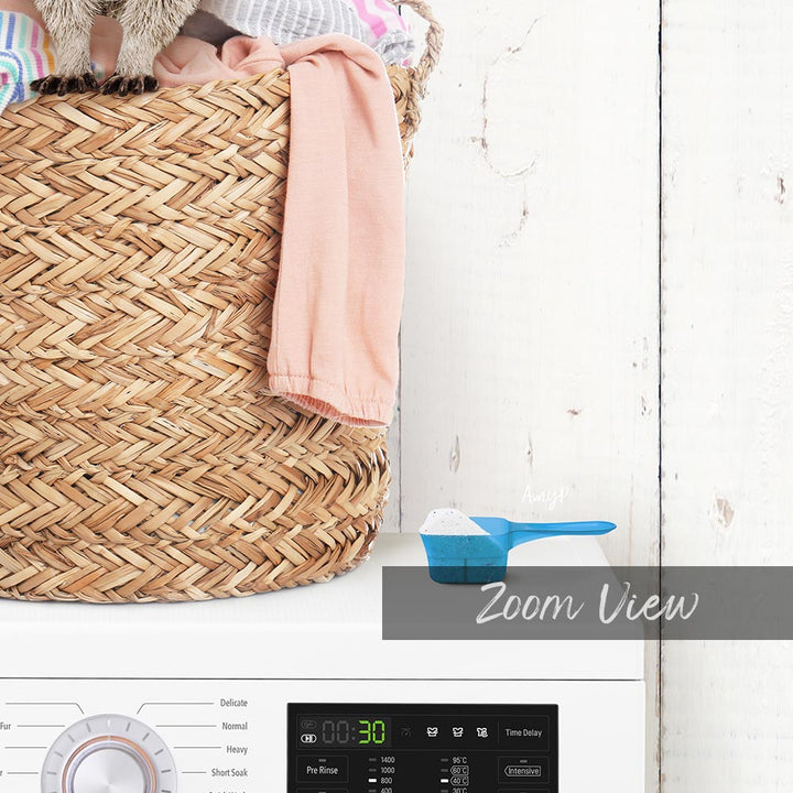 A woven laundry basket filled with clothes sits next to a white washing machine with a blue scoop.