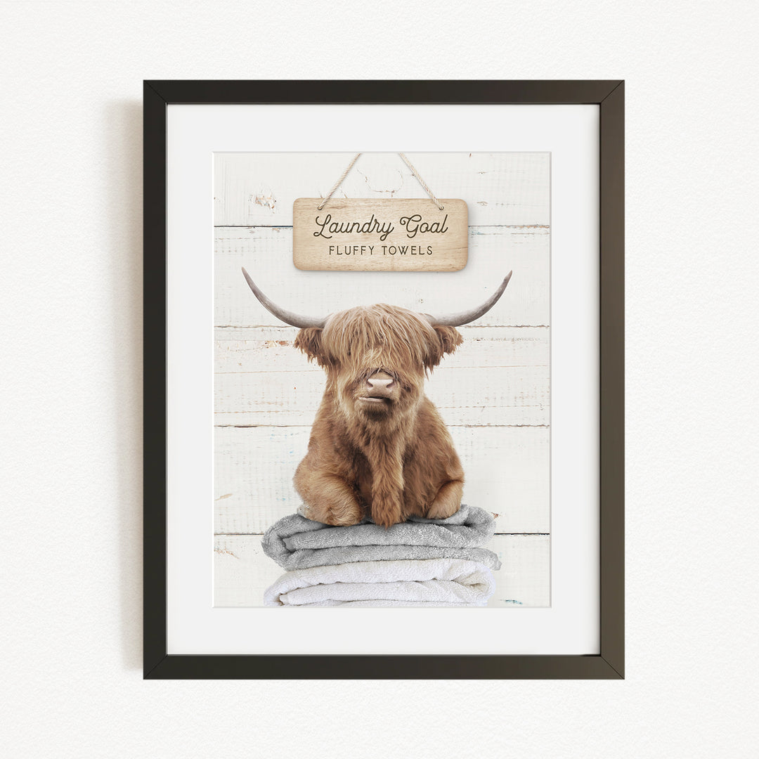 A framed image of a brown cow with long horns sitting on a stack of fluffy towels, with a sign above that says "Laundry Coal Fluffy Towels".