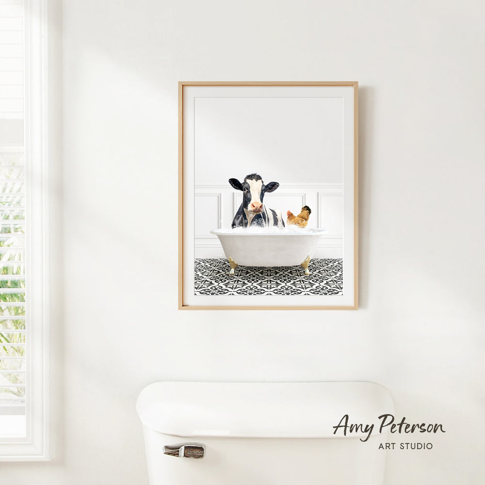 A framed image of a cow sitting in a bathtub, with a chicken in the tub, is hanging on a white wall.