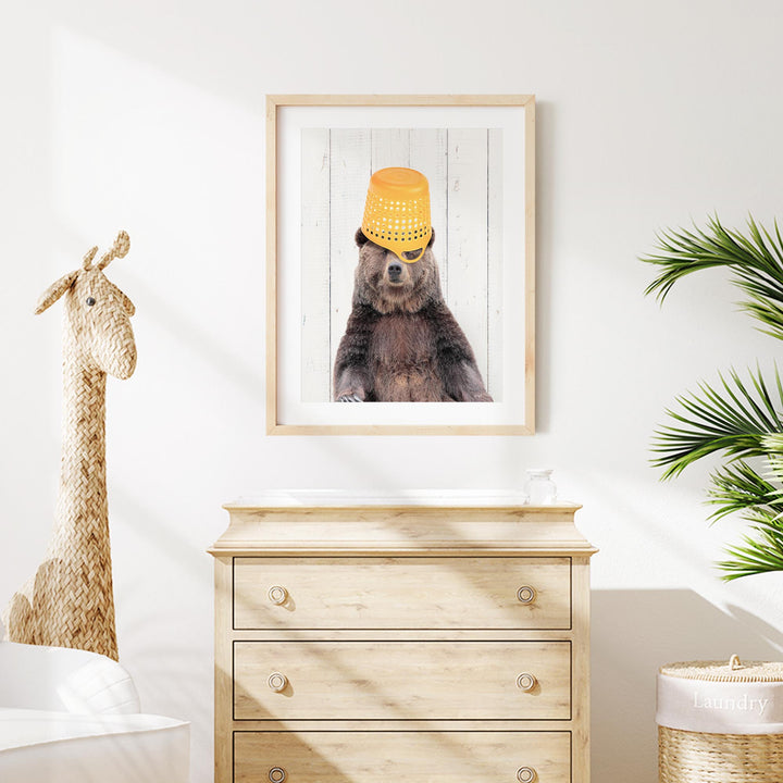 a wooden dresser with a framed picture of a bear wearing a yellow bucket hat on its head, placed above it. The dresser is situated in a room with a giraffe figurine and a potted plant.