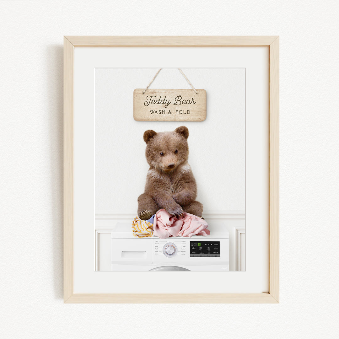 A framed image of a teddy bear sitting on a washing machine, with a sign above it that says "Teddy Bear Wash & Fold".
