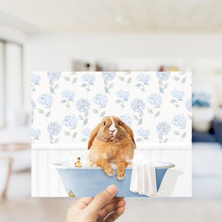 A hand holding a printed image of a brown rabbit sitting in a blue bathtub filled with water, surrounded by a floral pattern.
