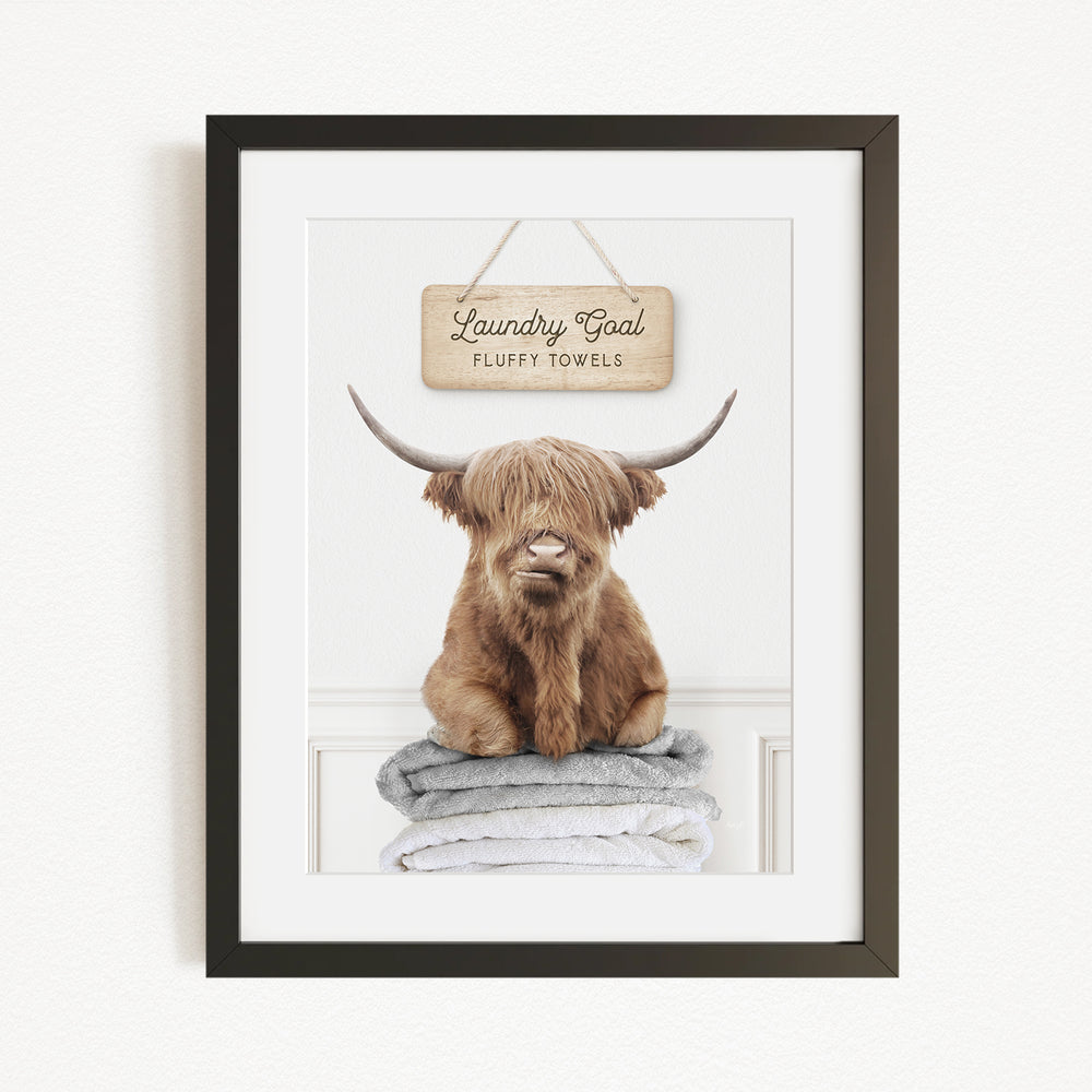 A framed image of a brown cow with long horns sitting on a stack of fluffy towels, with a sign above that says "Laundry Coal Fluffy Towels".