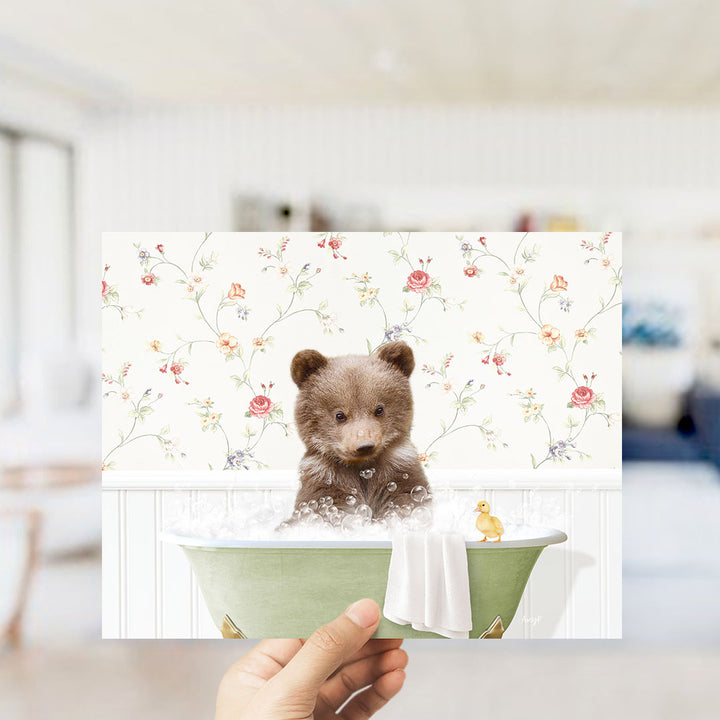 A hand holding a framed image of a small brown bear sitting in a bathtub filled with bubbles, with a yellow duck floating nearby.