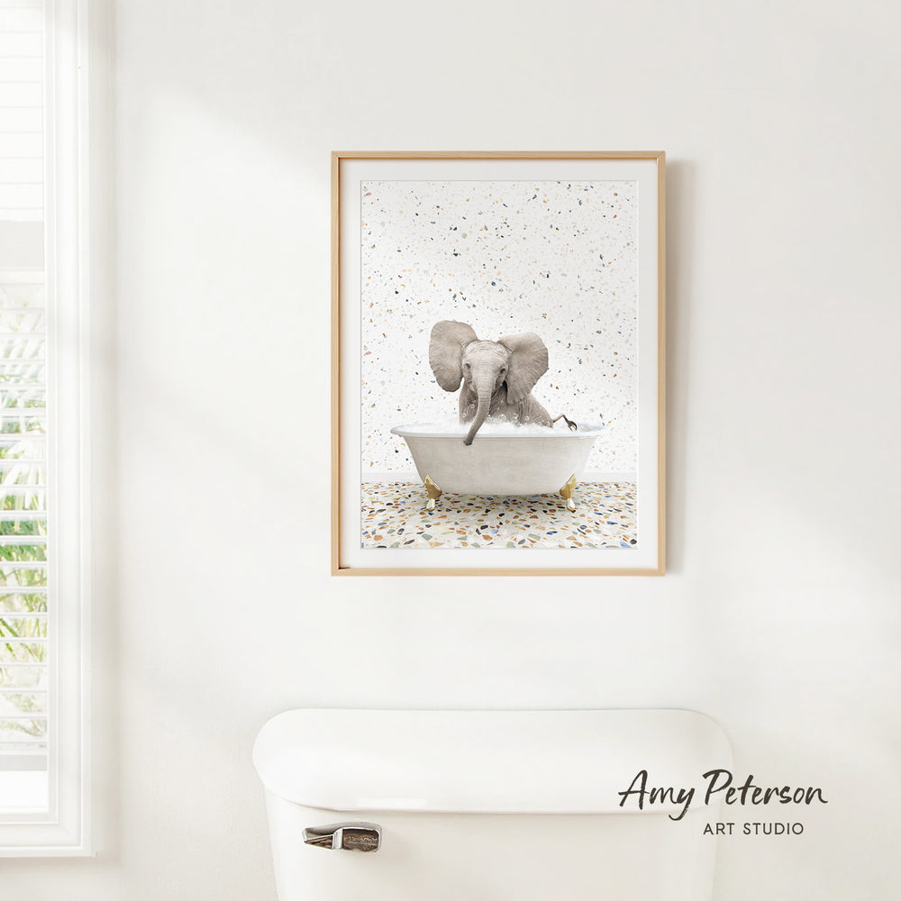 A framed print of an elephant sitting in a bathtub is hanging on a white wall.