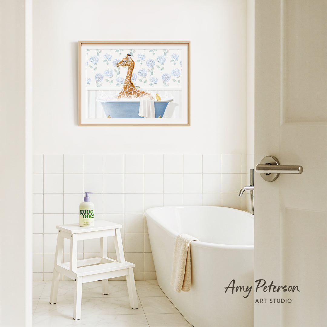 A framed painting of a giraffe sitting in a bathtub is hanging on a white tiled wall above a white bathtub in a bathroom.