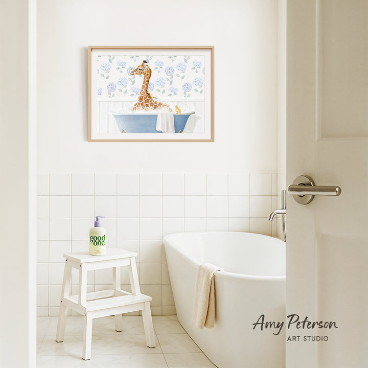 A framed painting of a giraffe sitting in a bathtub is hanging on a white tiled wall above a white bathtub in a bathroom.
