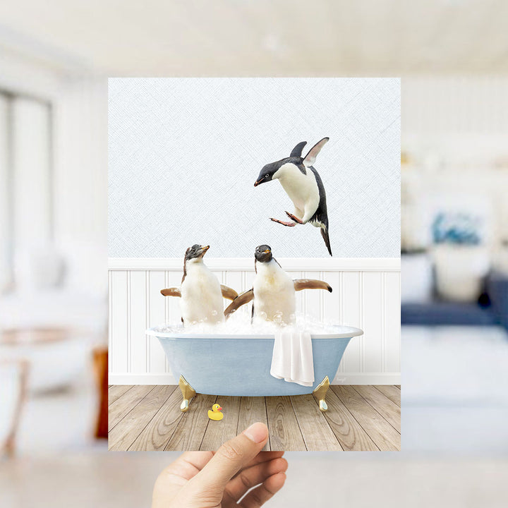 A hand holds up a framed image depicting two penguins sitting in a bathtub, with one penguin flying towards the other.