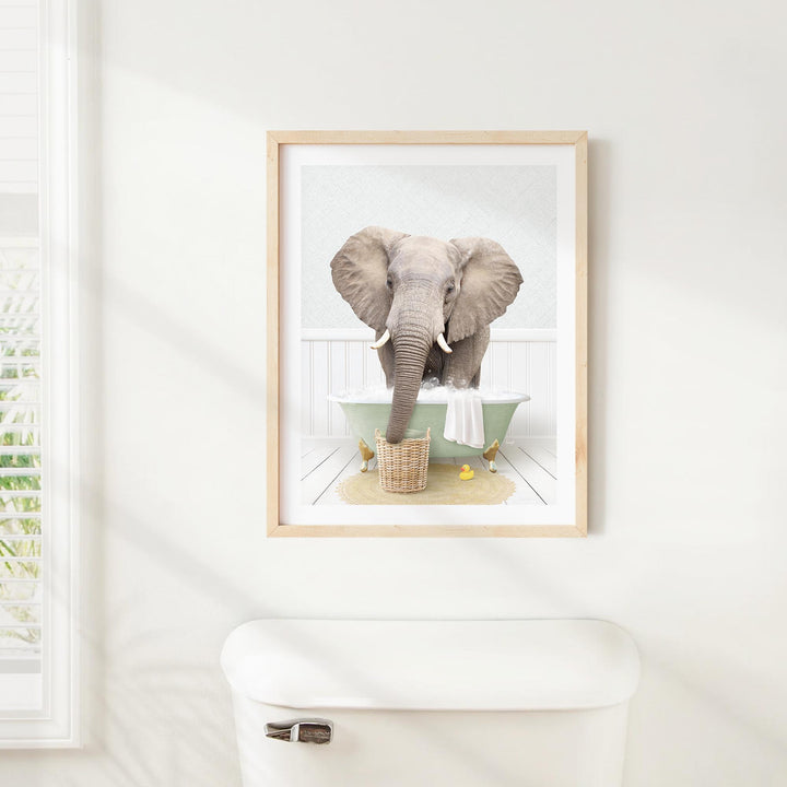 A framed image of an elephant standing in a bathtub, with a towel and basket of items nearby.