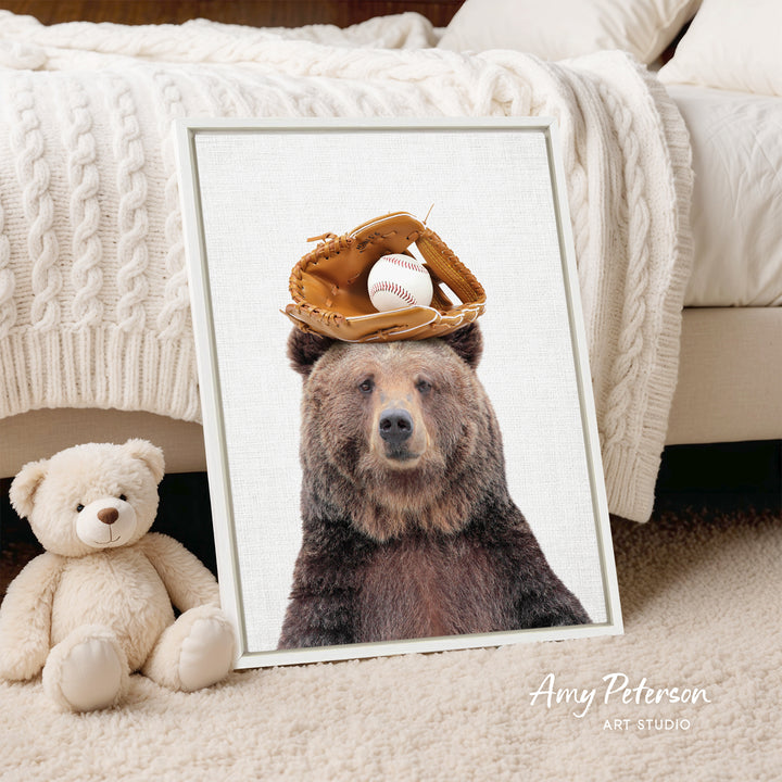 A framed image of a brown bear wearing a baseball glove on its head, with a teddy bear next to it, is displayed on a bed.