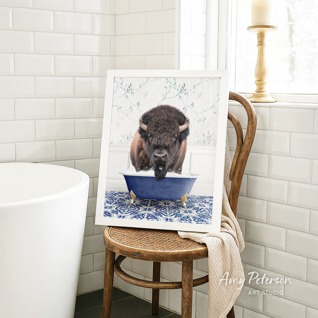 A framed image of a buffalo sitting in a blue bathtub, with a towel draped over the back of a wooden chair in a bathroom setting.