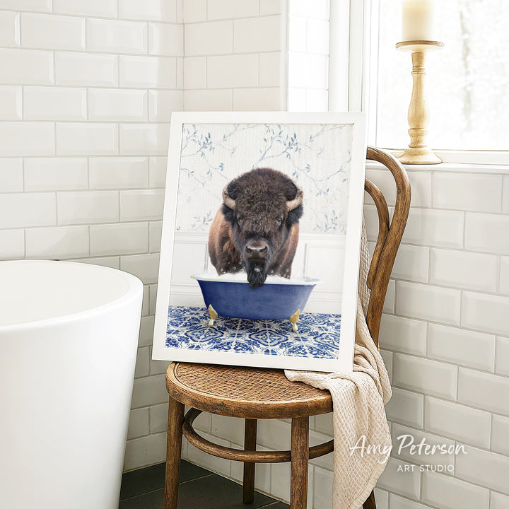 A framed image of a buffalo sitting in a blue bathtub, with a towel draped over the back of a wooden chair in a bathroom setting.