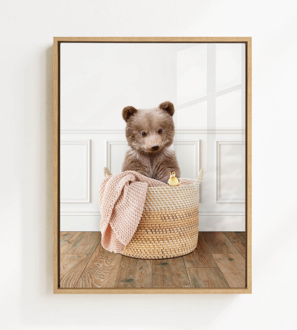 A brown bear sitting in a woven basket, with a pink blanket and a yellow duck toy nearby, against a white wall.